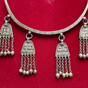 May include: A silver necklace with a circular band and three pendant charms. Each charm is a silver teardrop shape with a decorative pattern and a fringe of silver chains. The necklace is on a red background.