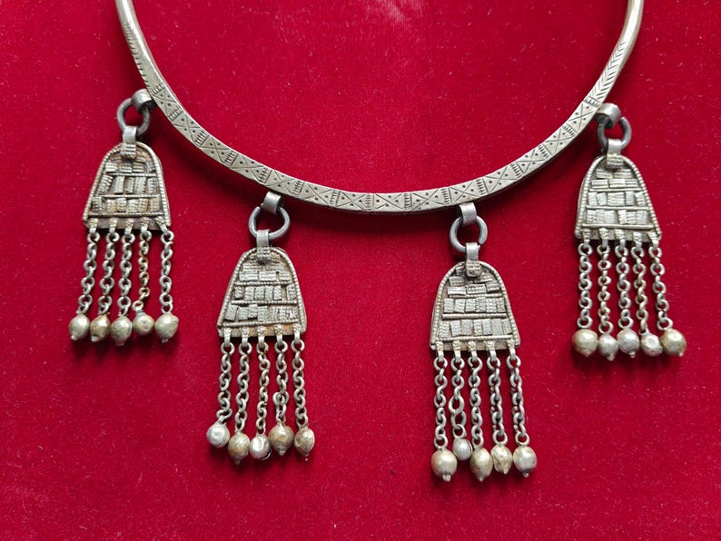 May include: A silver necklace with a circular band and three pendant charms. Each charm is a silver teardrop shape with a decorative pattern and a fringe of silver chains. The necklace is on a red background.