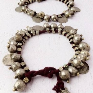 Può includere: Due cavigliere argentate con ciondoli a forma di monete e campanelle. Le cavigliere sono infilate su un cordoncino rosso scuro. I ciondoli sono di varie dimensioni e forme, e le monete sono piatte e rotonde. Presentate su una superficie bianca.