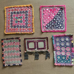 May include: Five embroidered fabric squares featuring colourful thread and small round mirrors. The squares are in various shades of pink, blue, green, and yellow.