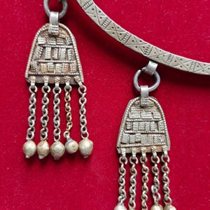 May include: A silver necklace with two pendants. Each pendant has a geometric design and is attached to a chain with small silver beads.