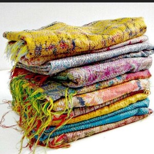 May include: A stack of colorful, patterned textiles. The textiles are in shades of yellow, blue, pink, and orange, with visible stitching and fringed edges. The fabrics appear to be layered, creating a textured and visually appealing display.