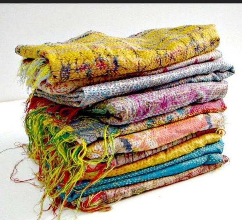 May include: A stack of colorful, patterned textiles. The textiles are in shades of yellow, blue, pink, and orange, with visible stitching and fringed edges. The fabrics appear to be layered, creating a textured and visually appealing display.