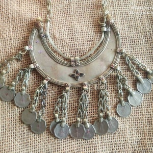 Puede incluir: Un collar de plata con un colgante de media luna y colgantes de monedas. El colgante está decorado con una pequeña piedra roja oscura.