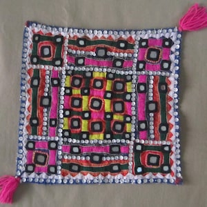 Peut inclure: Un coussin carré coloré avec un motif géométrique de tissu brodé et de petits miroirs. Le coussin a une frange rose sur deux coins.