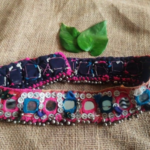 Puede incluir: Un cinturón de tela rosa y azul con pequeños espejos redondos y cuentas plateadas. El cinturón está decorado con bordados coloridos y tiene un respaldo de tela azul oscuro.