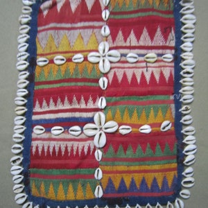 Puede incluir: Un textil cuadrado con un patrón geométrico en rojo, blanco, verde, amarillo y azul. El textil está bordeado con una fila de conchas de cauri blancas.