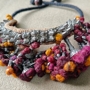 Op de afbeelding: Close-up van een handgemaakte ketting met een donkerrood en oranje kralenontwerp met zilverkleurige metalen accenten. De ketting heeft een geweven basis en een donkerblauw koord met een houten kraal.