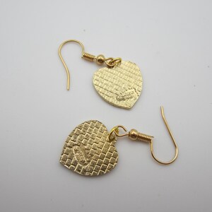 Puede incluir: Un par de pendientes dorados en forma de coraz&oacute;n. Los pendientes tienen una superficie texturizada y un cierre de gancho. Los colgantes de coraz&oacute;n est&aacute;n conectados a los ganchos con un peque&ntilde;o lazo dorado.
