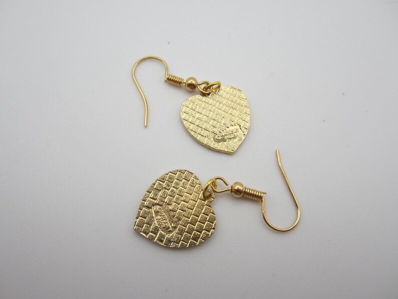Puede incluir: Un par de pendientes dorados en forma de coraz&oacute;n. Los pendientes tienen una superficie texturizada y un cierre de gancho. Los colgantes de coraz&oacute;n est&aacute;n conectados a los ganchos con un peque&ntilde;o lazo dorado.