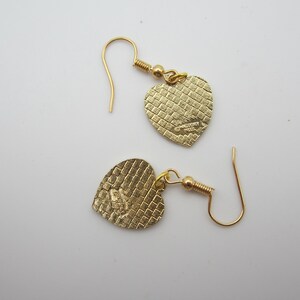 Puede incluir: Un par de pendientes dorados en forma de coraz&oacute;n. Los pendientes tienen una superficie texturizada y un cierre de gancho. Los colgantes de coraz&oacute;n est&aacute;n conectados a los ganchos con un peque&ntilde;o lazo dorado. Los pendientes se muestran sobre una superficie blanca.