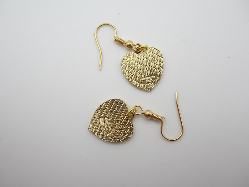 Puede incluir: Un par de pendientes dorados en forma de coraz&oacute;n. Los pendientes tienen una superficie texturizada y un cierre de gancho. Los colgantes de coraz&oacute;n est&aacute;n conectados a los ganchos con un peque&ntilde;o lazo dorado. Los pendientes se muestran sobre una superficie blanca.