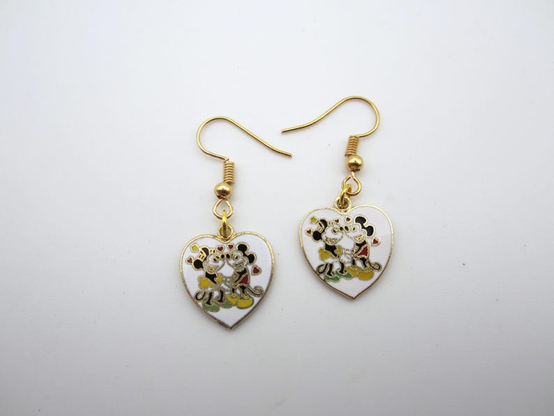Puede incluir: Un par de pendientes colgantes dorados. Cada pendiente presenta un dije en forma de coraz&oacute;n blanco con un dise&ntilde;o de pareja de dibujos animados en negro, rojo, amarillo y verde. Los pendientes tienen un cierre de gancho cl&aacute;sico.
