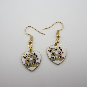 Puede incluir: Un par de pendientes dorados en forma de coraz&oacute;n. Cada pendiente presenta un coraz&oacute;n de esmalte blanco con un dise&ntilde;o de pareja de dibujos animados. La pareja de dibujos animados es de color negro, rojo, amarillo y verde. Los pendientes tienen cierres de gancho dorados.