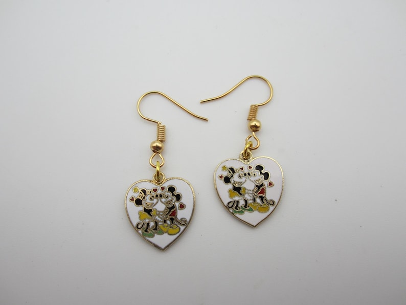 Puede incluir: Un par de pendientes dorados en forma de coraz&oacute;n. Cada pendiente presenta un coraz&oacute;n de esmalte blanco con un dise&ntilde;o de pareja de dibujos animados. La pareja de dibujos animados es de color negro, rojo, amarillo y verde. Los pendientes tienen cierres de gancho dorados.