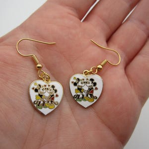 Puede incluir: Un par de pendientes en forma de coraz&oacute;n con ganchos dorados. Cada pendiente blanco presenta una pareja de dibujos animados en negro, rojo, amarillo y verde. Los pendientes se sostienen en una mano.