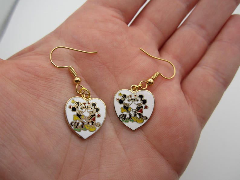 Puede incluir: Un par de pendientes en forma de coraz&oacute;n con ganchos dorados. Cada pendiente blanco presenta una pareja de dibujos animados en negro, rojo, amarillo y verde. Los pendientes se sostienen en una mano.