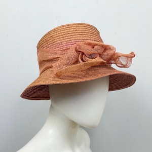 Puede incluir: Sombrero de paja marrón claro con ala ancha y una flor decorativa melocotón de malla. El sombrero se muestra sobre una cabeza de maniquí. Un accesorio elegante para los días soleados.