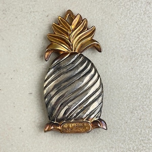 May include: A vintage Lenox pineapple brooch. The brooch features a gold-toned crown and base, with a silver-toned, textured body. The brooch has a detailed design and is a collectible item.