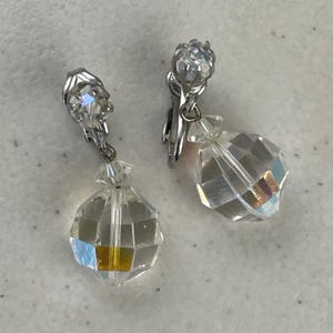 May include: A pair of vintage clip-on earrings featuring faceted crystal teardrop pendants. The earrings have silver-toned metal clips and small crystal accents at the top. The crystals reflect light, creating a sparkling effect.