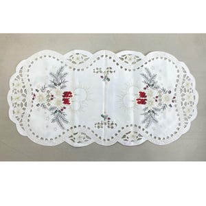 Könnte beinhalten: Weißer ovaler Tischläufer mit gewelltem Rand und gesticktem weihnachtlichem Blumendesign. Das Design umfasst weiße Poinsettien, rote Beeren und grüne Tannennadeln. Der Läufer ist ca. 91 cm lang.