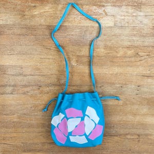 Può includere: Una borsa a tracolla turchese con cinturino abbinato. La borsa presenta un motivo di forme astratte rosa e bianche. La borsa è realizzata in un materiale liscio e ha una chiusura con coulisse.