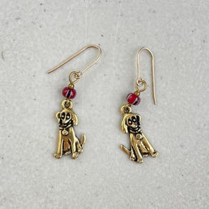 May include: A pair of gold-colored dog earrings. Each earring features a small, seated dog charm with a red and black beaded accent. The earrings have a simple hook design.