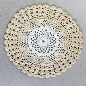 May include: Circular cream-coloured doily with a scalloped edge. The centre features a detailed, openwork design, transitioning to a more open pattern towards the outer edge. The doily is made of a crocheted material.