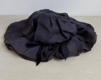 Puede incluir: Un sombrero de color gris carbón oscuro con una copa fruncida y plisada y un ala ancha, suave y con volantes. El sombrero está adornado con un pequeño lazo de cinta de terciopelo oscuro colocado en la parte delantera, añadiendo textura.