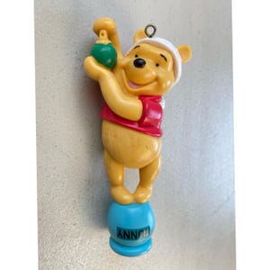 May include: A yellow Winnie the Pooh ornament holding a green ornament. Pooh is wearing a red shirt and a white hat. The ornament is standing on a blue base with the word "HUNNY" printed on it.