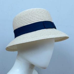 May include: A cream-colored straw hat with a dark blue ribbon band. The hat has a rounded crown and a wide brim, suitable for sun protection. The ribbon is tied in a bow on the side. The hat is displayed on a mannequin head.