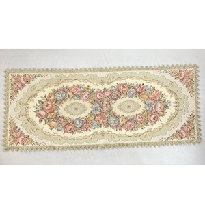 May include: Cream-colored floral table runner with an oval floral design in the center. The design features pink, blue, and yellow flowers. The edges are trimmed with a scalloped border and gold fringe.