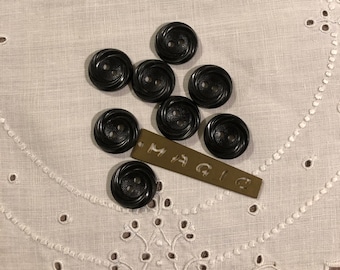 May include: Eight black plastic buttons with two holes each. The buttons have a raised, twisted design. The buttons are arranged in a loose circle on a white lace tablecloth. A small brass tag with the word "MAGIC" is visible in the lower right corner of the image.