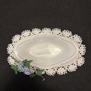 May include: White oval ceramic dish with blue lettering that reads "The Good Fellows Club". The dish is on a white crocheted doily with a sprig of blue flowers.
