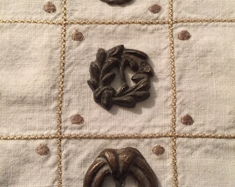 Könnte beinhalten: Drei antike bronzefarbene Metallknöpfe mit komplizierten Designs. Der obere Knopf ist ein kreisförmiges Design mit einer erhabenen Mitte. Der mittlere Knopf ist ein Kranzdesign. Der untere Knopf ist ein ovales Design mit einer erhabenen Mitte.
