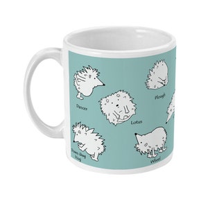 May include: White ceramic mug with a light teal background featuring cartoon hedgehogs in yoga poses. Poses include Dancer, Lotus, Plough, and Wheel. The mug has a white handle and is suitable for hot or cold beverages.