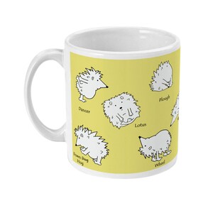 May include: White ceramic mug with a yellow background featuring cartoon hedgehogs in various yoga poses. The mug has a white handle and the poses are labeled "Dancer", "Lotus", "Plough", "Wheel", and "Down Dog Hog".
