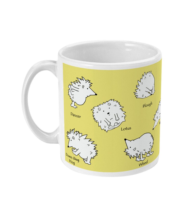 May include: White ceramic mug with a yellow background featuring cartoon hedgehogs in various yoga poses. The mug has a white handle and the poses are labeled "Dancer", "Lotus", "Plough", "Wheel", and "Down Dog Hog".
