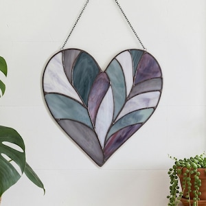 May include: A heart-shaped stained glass wall hanging with a chain for display. The glass is divided into sections of white, purple, gray, and teal. The artwork is hanging on a white wall, with a plant and a window in the background.