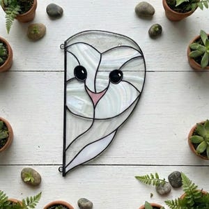May include: A stained glass suncatcher shaped like the left half of a barn owl face, featuring iridescent white glass panels, black solder lines, and a small pink glass beak. The owl has two dark circular eyes and a curved, symmetrical design.
