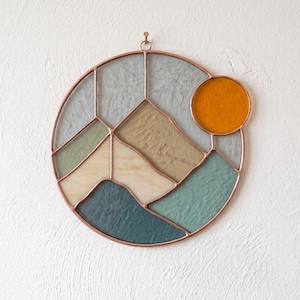 Puede incluir: Un atrapasoles circular de vidrieras que muestra un paisaje montañoso con un sol naranja brillante. La pieza está construida con líneas de cobre y contiene paneles de vidrio texturizado en tonos gris claro, beige, verde salvia y azul verdoso oscuro.