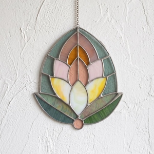 Op de afbeelding: Een glas-in-lood suncatcher in de vorm van een gestileerde lotusbloem. Het object bestaat uit segmenten van doorschijnend glas in saliegroen, zachtroze, amber en lichtgeel, bijeengehouden door dunne donkere metalen soldeerlijnen en een ketting.