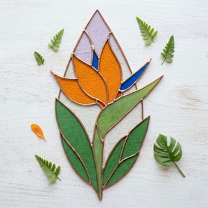 Puede incluir: Vidriera con forma de flor de ave del paraíso. La flor presenta vidrio naranja, azul y verde, delineado con cobre. Hojas de helecho verdes y una hoja de monstera se disponen alrededor de la obra sobre una superficie de madera blanca.
