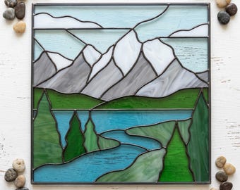 Peut inclure: Un panneau carré en vitrail représentant un paysage de montagne avec des sommets gris enneigés, une forêt vert vif et une rivière bleue. La scène est encadrée de métal sombre, avec des sections en verre clair pour le ciel.