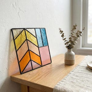 May include: A colorful stained glass panel with a black metal frame. The geometric design features sections of yellow, orange, pink, and blue glass. The panel is displayed on a wooden surface, with a vase of dried flowers in the background.