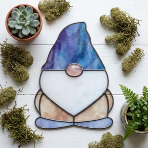 May include: A stained glass gnome with a blue and purple hat, white beard, and a pink stone nose. The gnome is surrounded by moss and small potted plants. The gnome is a decorative item.