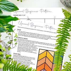 May include: Two sheets of paper with the text "Upward Beginner Pattern" and instructions for a stained glass project. The finished size is 8" x 8". The papers are surrounded by green plants.
