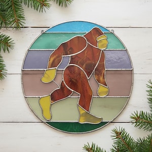 Könnte beinhalten: Runde Glasmalerei mit einem braunen Bigfoot in Bewegung. Das Design verwendet Gelb, Braun, Grün, Lila und Blau, mit einem silbernen Metallrahmen. Tannenzweige umrahmen das Panel.
