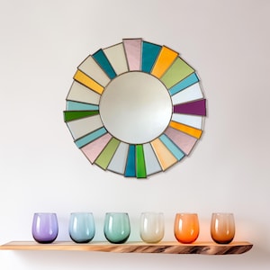 May include: A round mirror with a stained glass design in shades of pink, yellow, green, blue, and purple. The mirror is surrounded by a gold frame and is hanging on a white wall. There are six colorful glasses on a wooden shelf below the mirror.