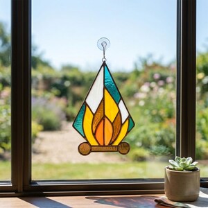 May include: A diamond-shaped stained glass suncatcher featuring a flame-like design in shades of orange, yellow, and teal, framed in dark metal solder. The piece is suspended by a clear suction cup hook against a window pane with a garden view.